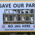 A yard sign reads 'Save Our Park, No Jail Here' with an image of rabbit statues behind bars overlaid on a park photo.