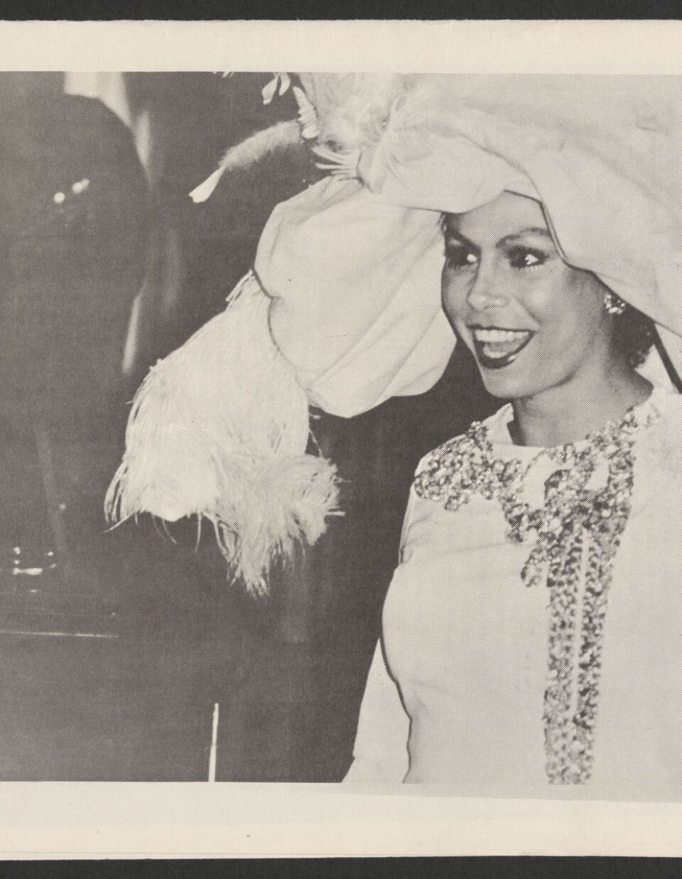 A black and white photo of a trans woman performer wearing a giant hat and sequined dress.