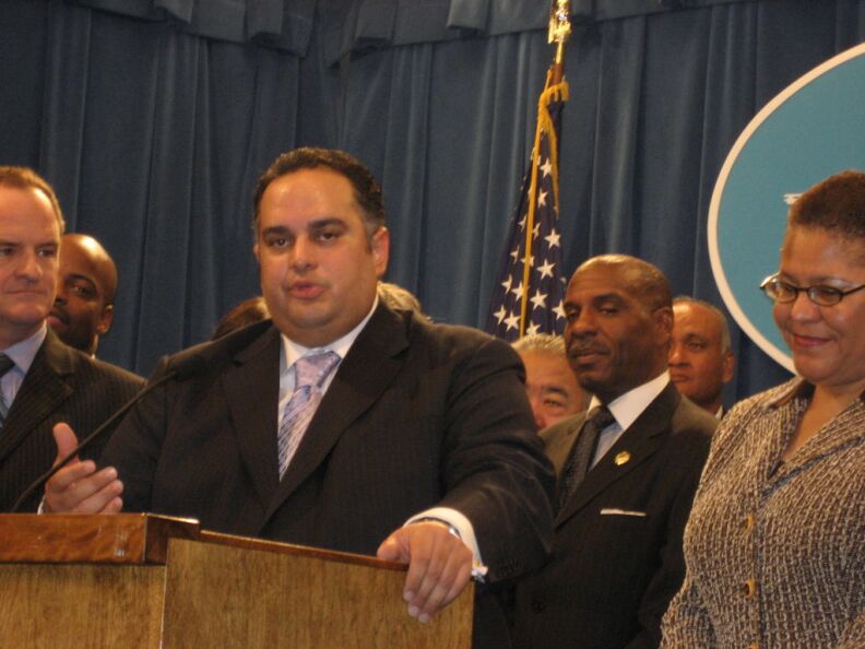 A man with medium-tone skin wears a dark suit with a light tie at a lectern. He's surrounded by a diverse group of people.