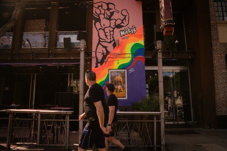 Two people with light skin tones walk down a sidewalk looking away from the camera. Next to them is art for Micky's, a gay club. It has an illustration of a muscular man and wavy rainbow pride colors.