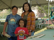 An older woman with dark hair stands with her young grandchildren outside under a shade structure. 
