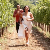 A female-presenting person in a white dress and a male-presenting person in an orange shirt running through a grove of young trees.