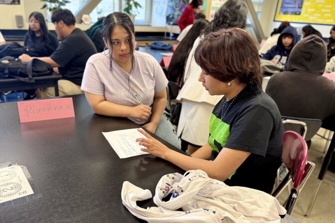 A young person with medium skin tone sits at a black table looking over a piece of paper. Next to them, a young woman with medium skin tone watches, with a sign next to her that says "Roxanna."