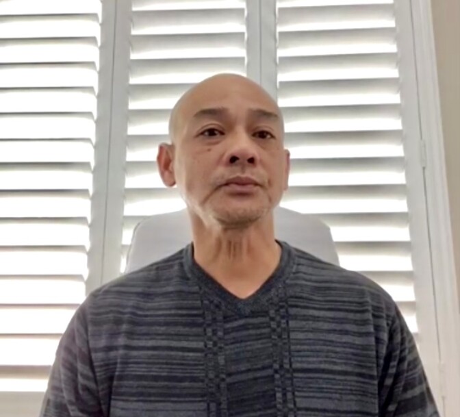 A 48-year-old man of Vietnamese descent with a shaven head looks somberly into the camera, with a window with slatted blinds behind him. 