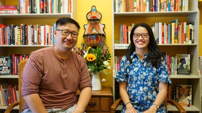 Two people with glasses stand in front of a few bookshelves lined up against a yellow wall.