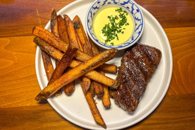 A neatly plated steak frites dish featuring a seared, medium-rare cut of steak with visible grill marks, thick-cut golden fries, and a small bowl of creamy yellow sauce garnished with chopped chives, all served on a white plate atop a warm wooden table.