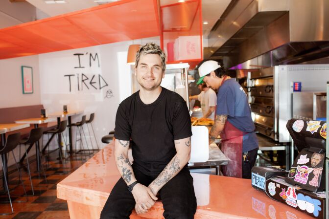 A light-skinned man wearing a black T-shirt and black pants sits on a polished light orange countertop with his hands in his lap. He has bleached blonde hair and blue eyes. Behind him, on his left, is a pizzeria space with the words "I'm Tired" on the back wall in black lettering. On his right is a man with light skin and a black mustache wearing a blue T-shirt and red apron with white flour. Behind him are silver metal pizza ovens. 