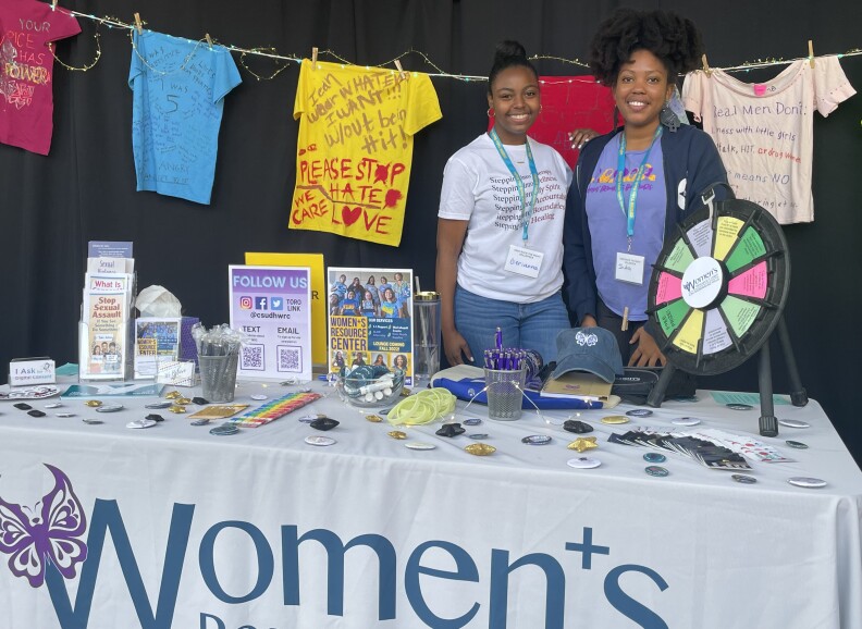 Two female students pose behind a table teeming with giveaways, including buttons, pens, and pamphlets. T-shirts of many colors hang behind them, with messages against sexual assault.