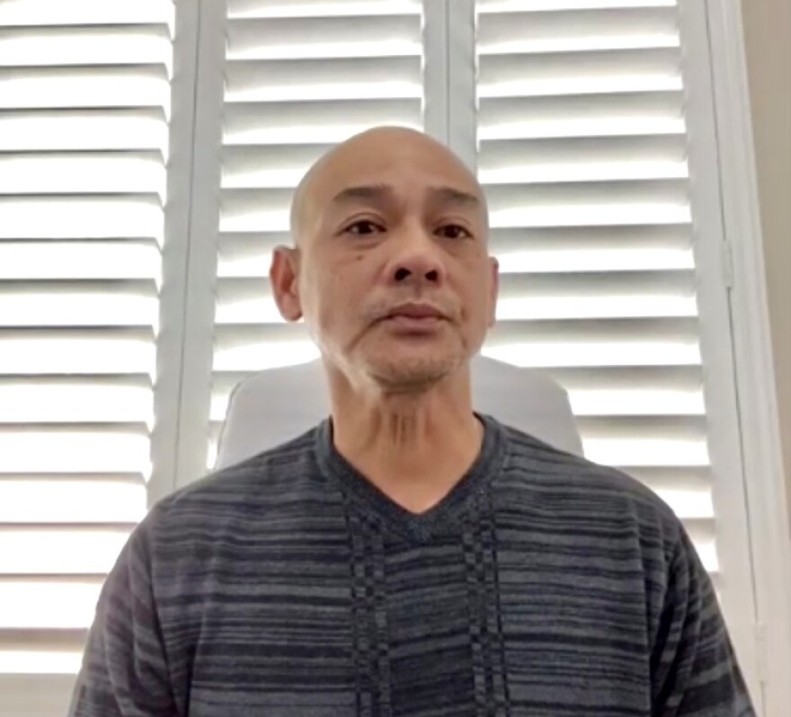 A 48-year-old man of Vietnamese descent with a shaven head looks somberly into the camera, with a window with slatted blinds behind him.