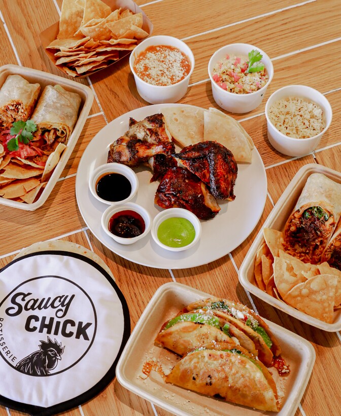 An image of a roasted red chicken cut into parts on a white plate with small containers with different colored sauces. Next to the chicken are two folded flour tortillas. Around the plate are three paper white containers containing beans, rice, and corn, Surrounding the plate are rectangular containers with burritos and tacos with a side of chips. 