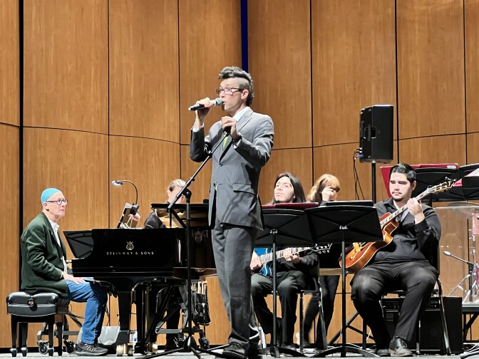 A man with a mohawk in a gray suit introduces the next song on stage with guitar and piano players behind him.