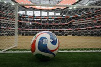 A multi-colored soccer ball sits on a patch of grass and beside a net. In the background is an empty stadium.