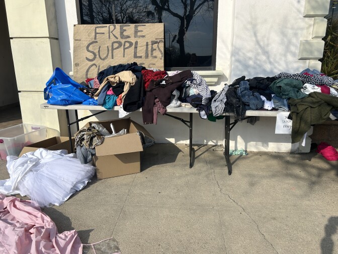 Clothes are piled on tables. A hand-written sign says "Free Supplies Clothes"