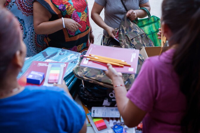 Hands sort through school supplies — notebooks, pencils and backpacks — on a table surrounded by several people.