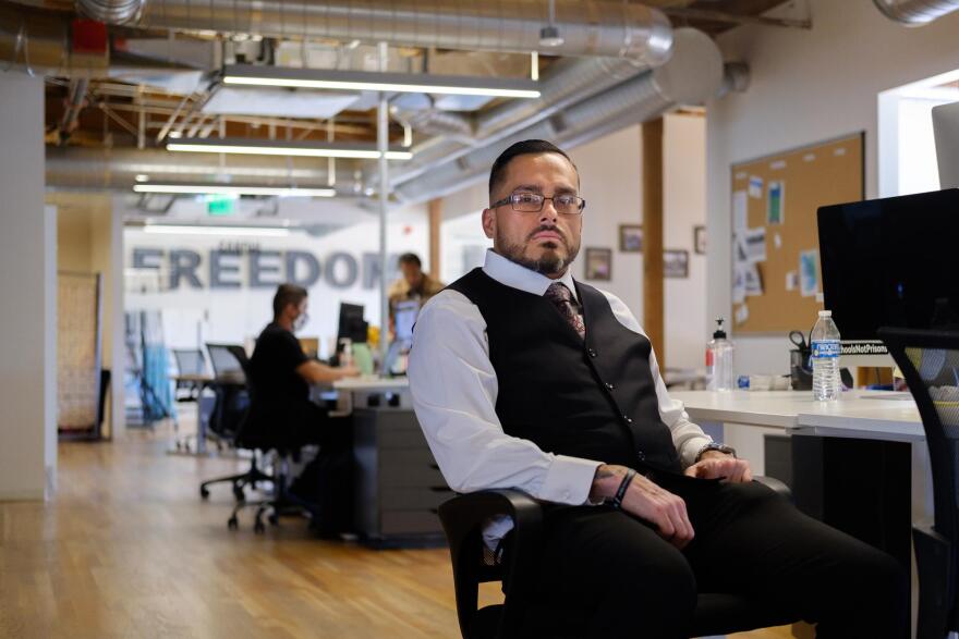 José Norrington sits at a desk facing the camera in the offices of the Anti-Recidivism Coalition. He's working a suit and tie with a vest. Two people can be seen in the background, and at the rear of the room the word "freedom" can be seen on the wall.