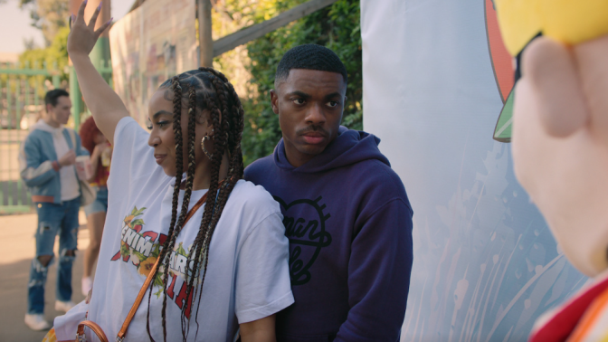 A woman waves with her hand up while a man stands close behind her.