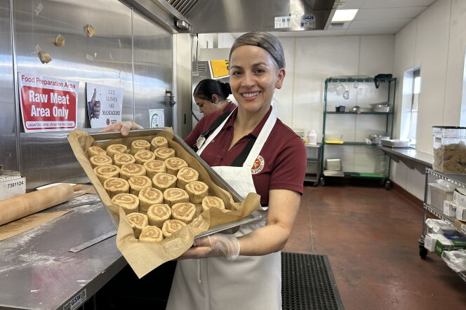 A woman wearing a red polo, apron and hair net holds a baking sheet with freshly rolled cinnamon rolls.