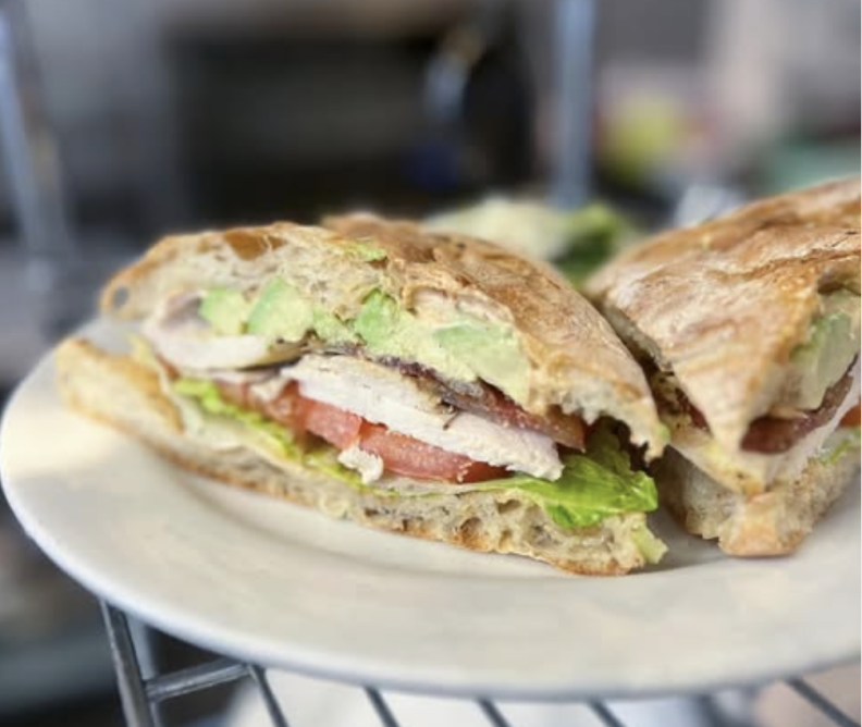 A sandwich cut in half showing the insides sits on a white plate. The crusty bread contains creamy avocado, slices of chicken, red tomatoes and green lettuce.