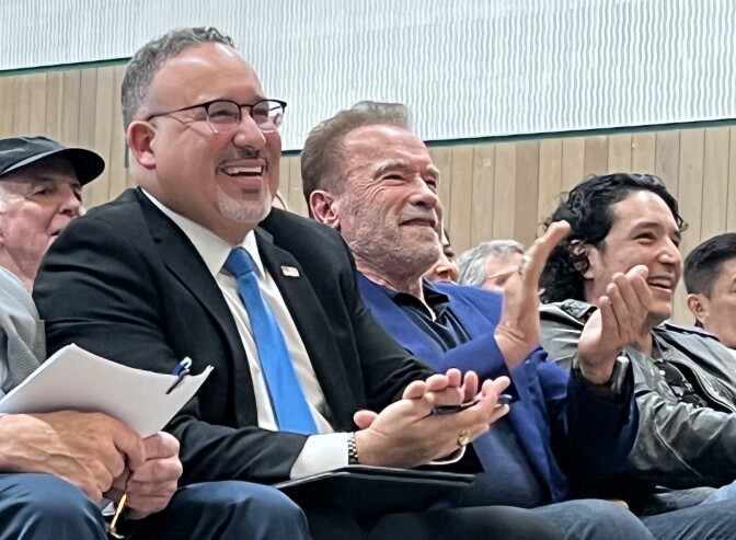 A light-skinned brown man with glasses and a blue tie and another man, who is white and wearing a blue suit, sit in a theater and applaud.