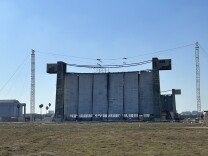  A view across a dirt and grass field at a wide, tall concrete-looking wall with six panels and concrete towers on either side. 