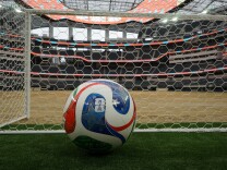 A multi-colored soccer ball sits on a patch of grass and beside a net. In the background is an empty stadium.