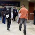 Adults and children wearing backpacks are seen from behind, walking toward the entrance of an elementary school. 