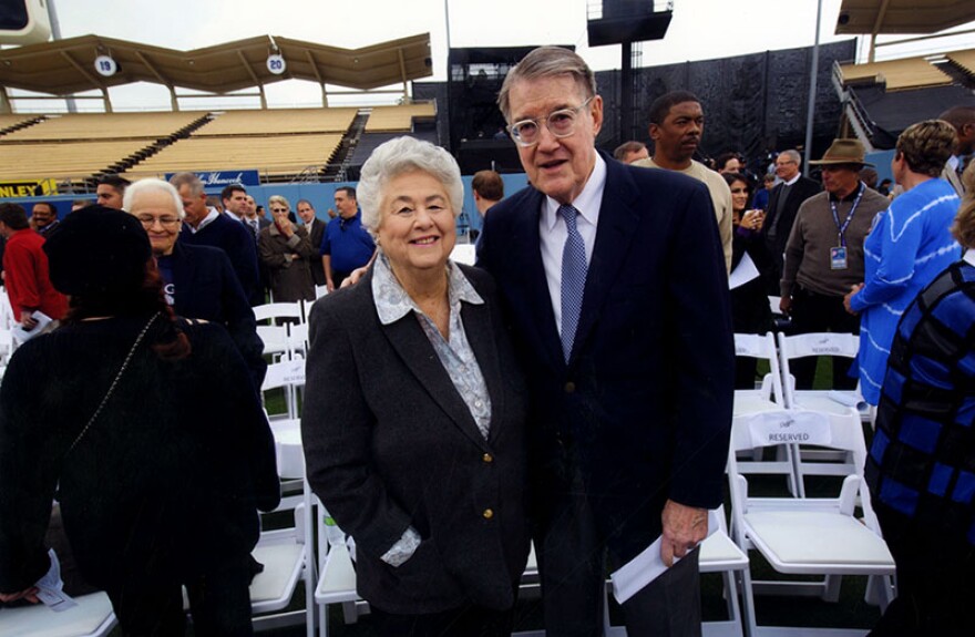 A grey-haired woman with a light skin tone wears a suit jacket and collared shirt. An older man in glasses and a suit has his arm around her on the field at Dodger Stadium