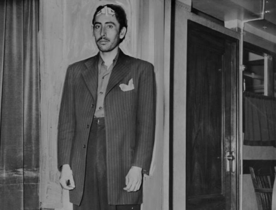 A man with medium-tone skin stands with his arms to his side in a suit with an oversized blazer and a pocket square.