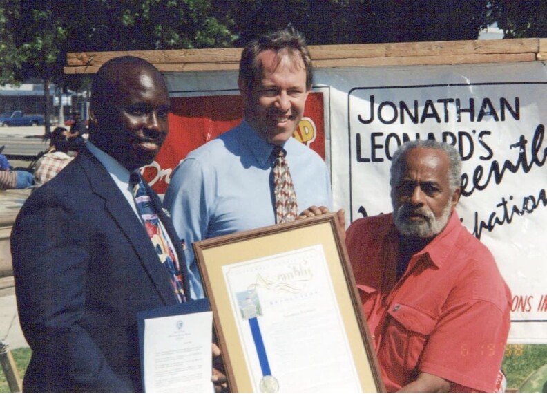 A dark-skinned Black man, a light-skinned white man and a medium-skinned Black man hold plaques in front of a white sign outside.