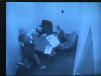Grainy security camera footage of the inside of an interrogation room. A blonde haired woman is standing next to a table, next to a man on the side who is sitting on a chair. Both are looking at a third man who has his head down and is looking at the table.