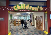 An entryway with a large yellow sign above that reads "Children's." Through the entry, there's a female-presenting person next to a baby stroller and shelves of books in the distance. 