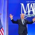 A man wearing a dark suit and tie holds his arms out. His mouth is open. Behind him is a sign against a blue background that reads, "MAHA Summit" and an American flag stands to his right