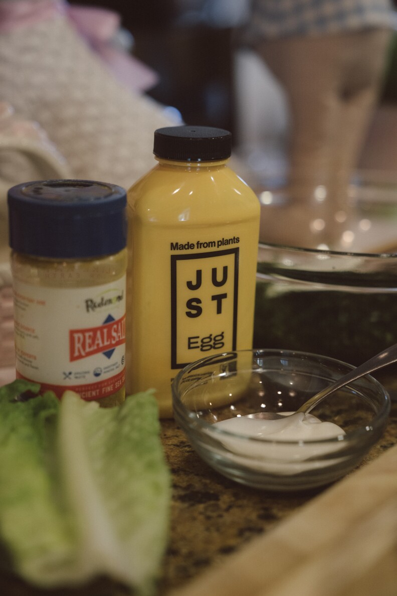 A yellow plastic bottle on a marble countertop that reads "JUST EGG" and "Made from plants." In front of the bottle there's a small glass bowl with yogurt and a piece of romaine lettuce.