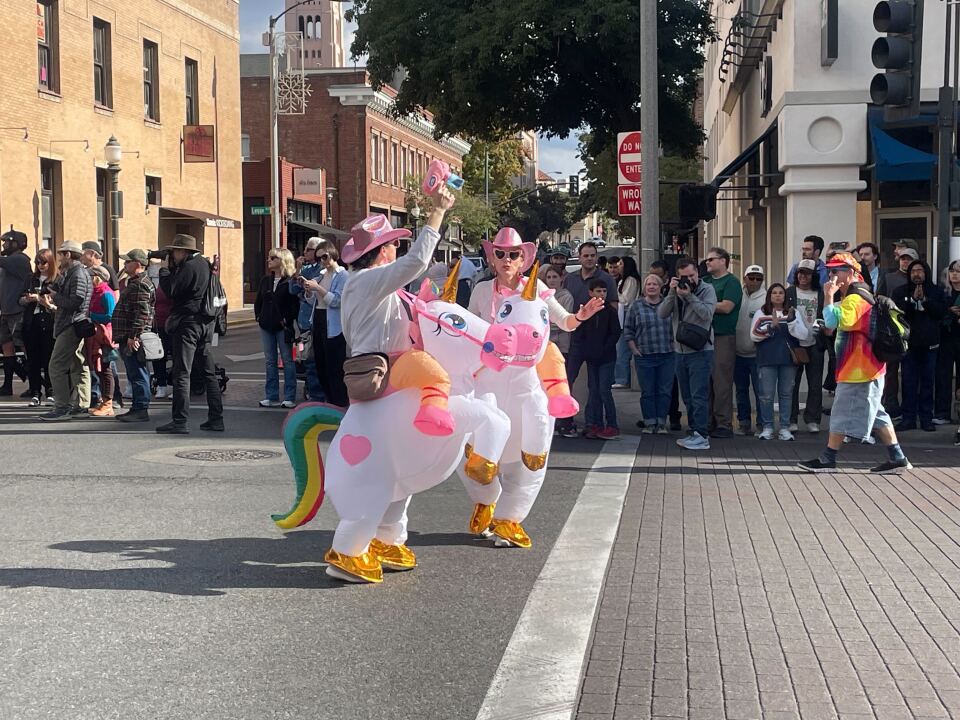 Two people dressed up in blow-up costumes that make it appear they are riding pink and white unicorns. The unicorns have rainbow tails. In the background, parade-goers take pictures.