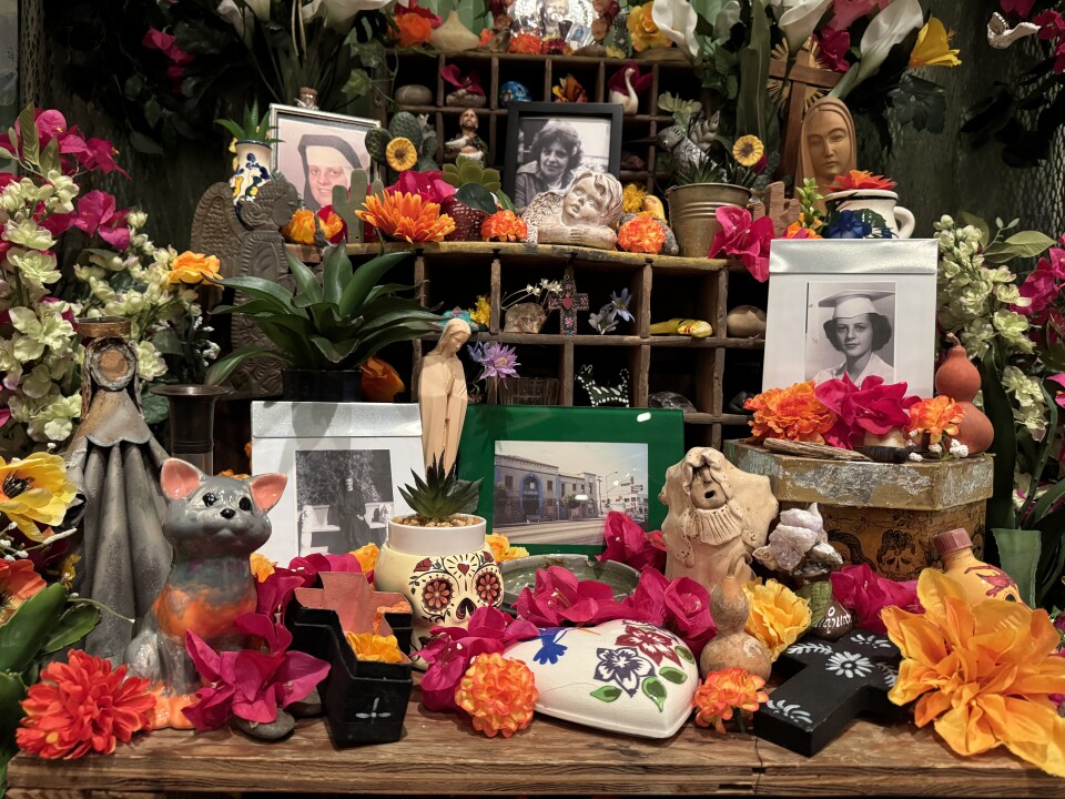 An altar features a myriad flowers, an orange cross, a black cross, a mouse-like animal and framed photos of a nun.