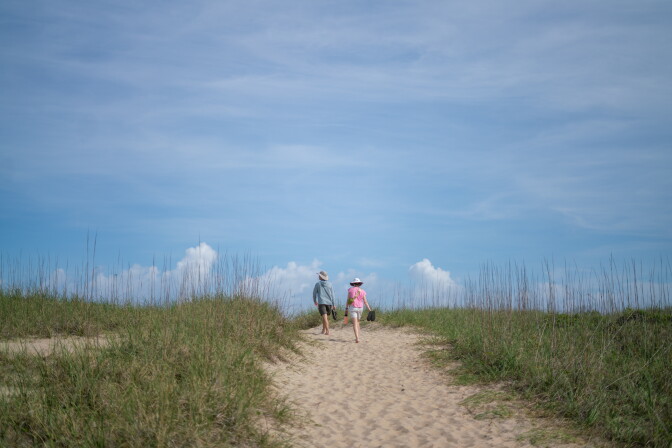 Two people walk along a sand path at a beach.