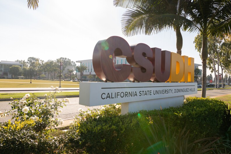 A landscape photo of a large red and yellow block letter sign that reads "CSUDH" below the block letters a white block with black letters reads "California State University, Dominguez Hills." Bushes surround the sign at the base and behind the sign, towards the right of frame there are two palm trees.