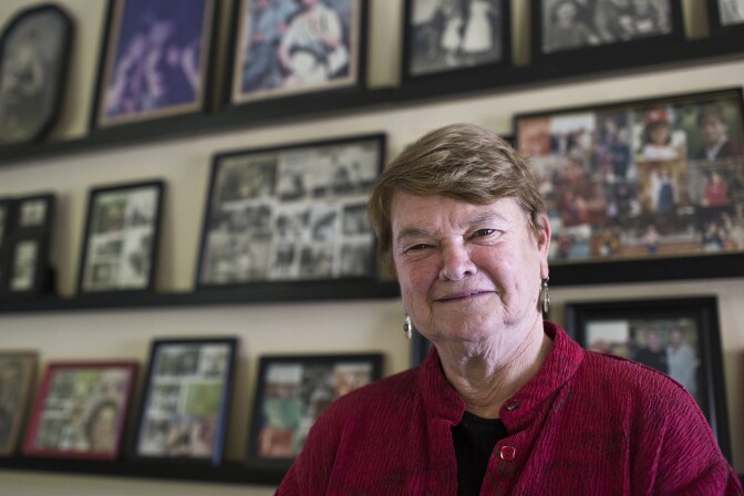 A white woman with short cropped, light-colored hair is shown wearing a red button-down shirt over a black top. She stands in front of three rows of framed photographs. The images are blurry in the distance. 