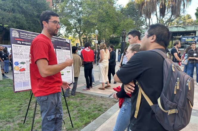 A man with light skin tone stands before a scientific research poster in a courtyard. In front of him, three other people listen as he speaks. In the background, people pause to look at other posters. 