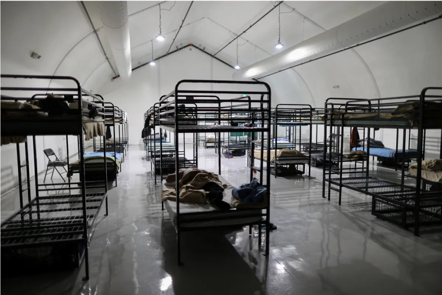 A row of metal bunk beds are housed within a white temporary structure with glossy concrete floors.