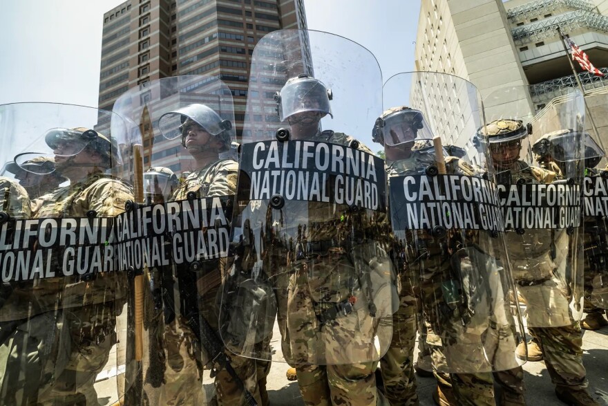 A row of California National Guard soldiers stand in a row, holding clear shields in front of them with the words "California National Guard." The soldiers are wearing camouflauge uniforms and helmets with face shields