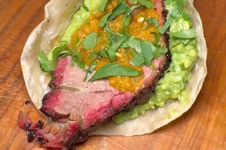 A hand wearing a black food prep glove holds a brisket taco with guacamole, salsa, and cilantro.