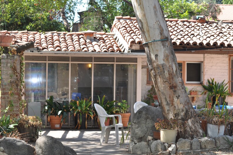 A one-story, white brick hose with Spanish-style tiles on the slanted roof. One wall is floor to ceiling windows. Outside the house are large potted plants, trees and several white plastic chairs. 