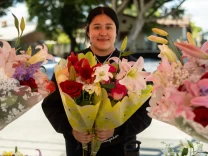 A young woman smiles as she holds two bouquets of flowers with more bouquets around her near a street.
