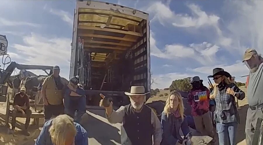 A male presenting person in a cowboy hat and vest points to the left, while surrounded by a film crew with an open  cargo truck in the background. 