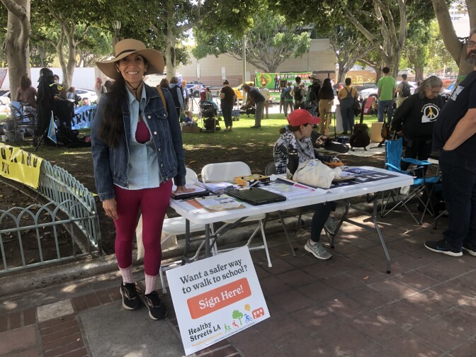 A woman wearing a sun hat, jean jacket and red pants smiles and stands next to a table with flyers. Groups of people are in the background. 
