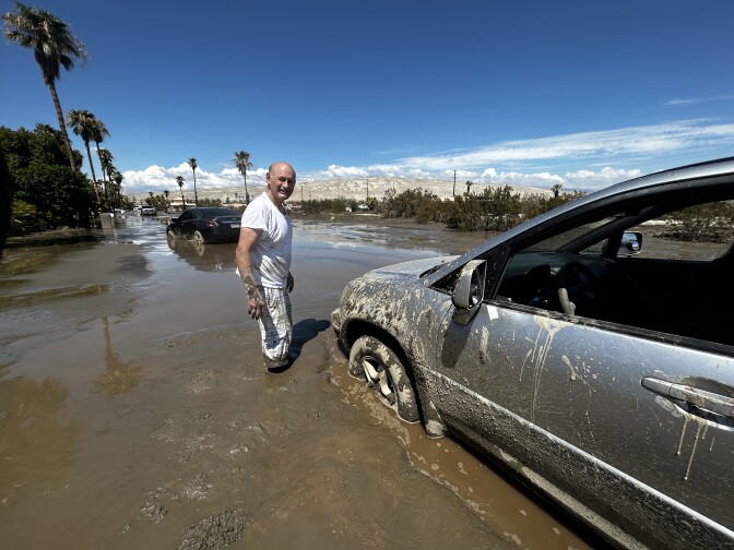 A middle aged, balding, light skinned man wearing a white t-shirt and mud-splatted white pants stands knee deep in mud beside a car stuck in the mud