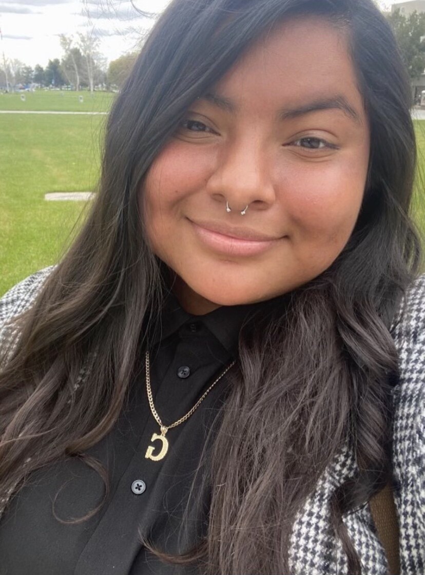 A selfie of a young woman with medium-brown skin and dark brown hair. She has a small silver nose ring and wears a golden necklace and black shirt. 