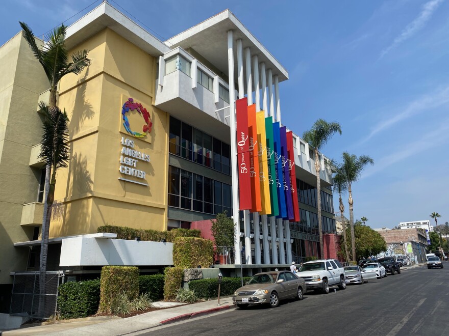 An exterior shot of the LA LGBT Center in Hollywood. The building features long, thin white columns that hold a rainbow of colored banners.