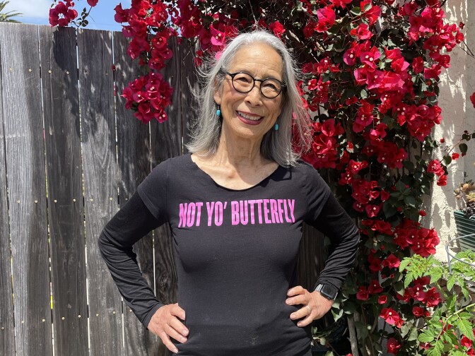 An 84-year-old Japanese American woman wears shoulder-length gray hair and round black glasses as she poses in front of a fuchsia bougainvillea.